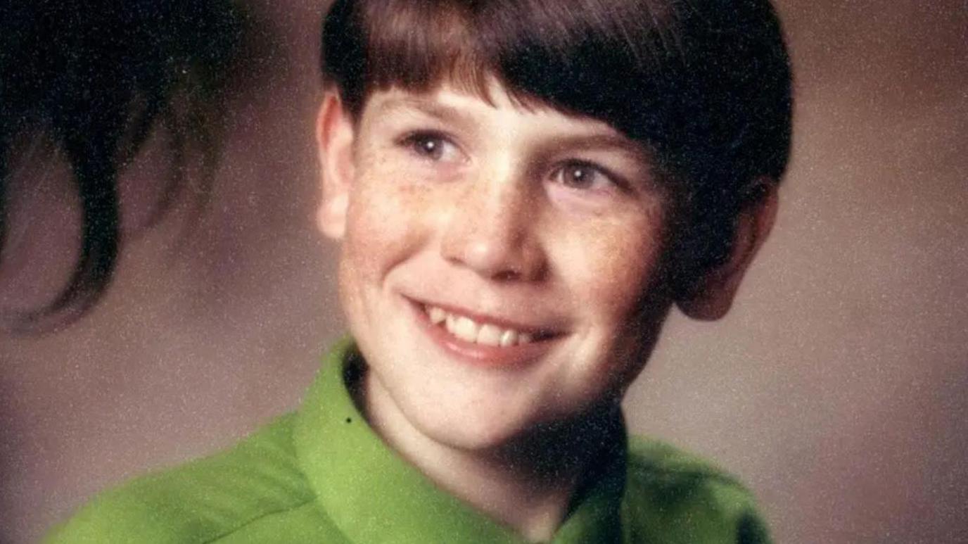 A young boy with brown hair smiles at something off-camera. He is wearing a bright green shirt. 