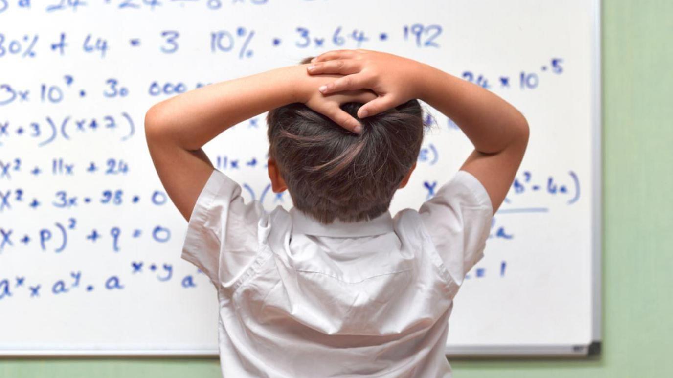 A child with brown hair and a white shirt with his back to the camera, looking at a white board with a lot of mathematical equations on it