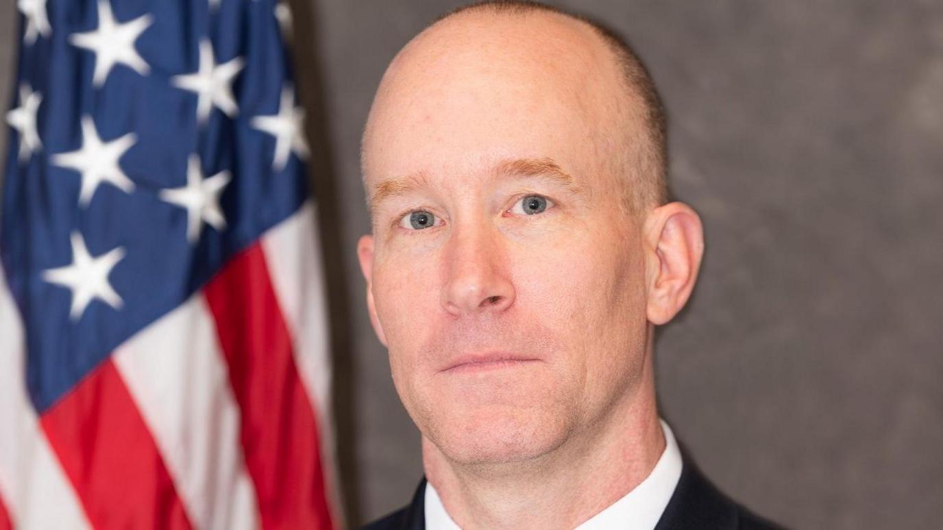 A bald man, pictured from the shoulders up. There is a US flag in the background. 