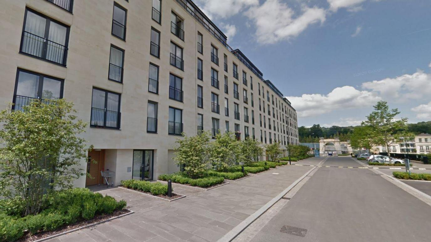 A block of modern flats on a wide road in Bath. The building is five storeys with a sandstone coloured frontage and black framed windows and Juliet balconies.