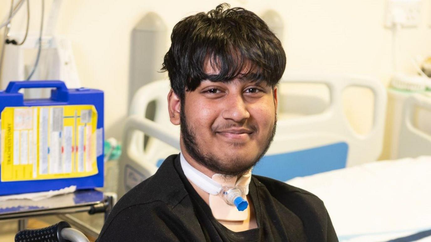 Ayyub is in his hospital room. There is a bed in the background and he is smiling at the camera with his tracheostomy tube in view