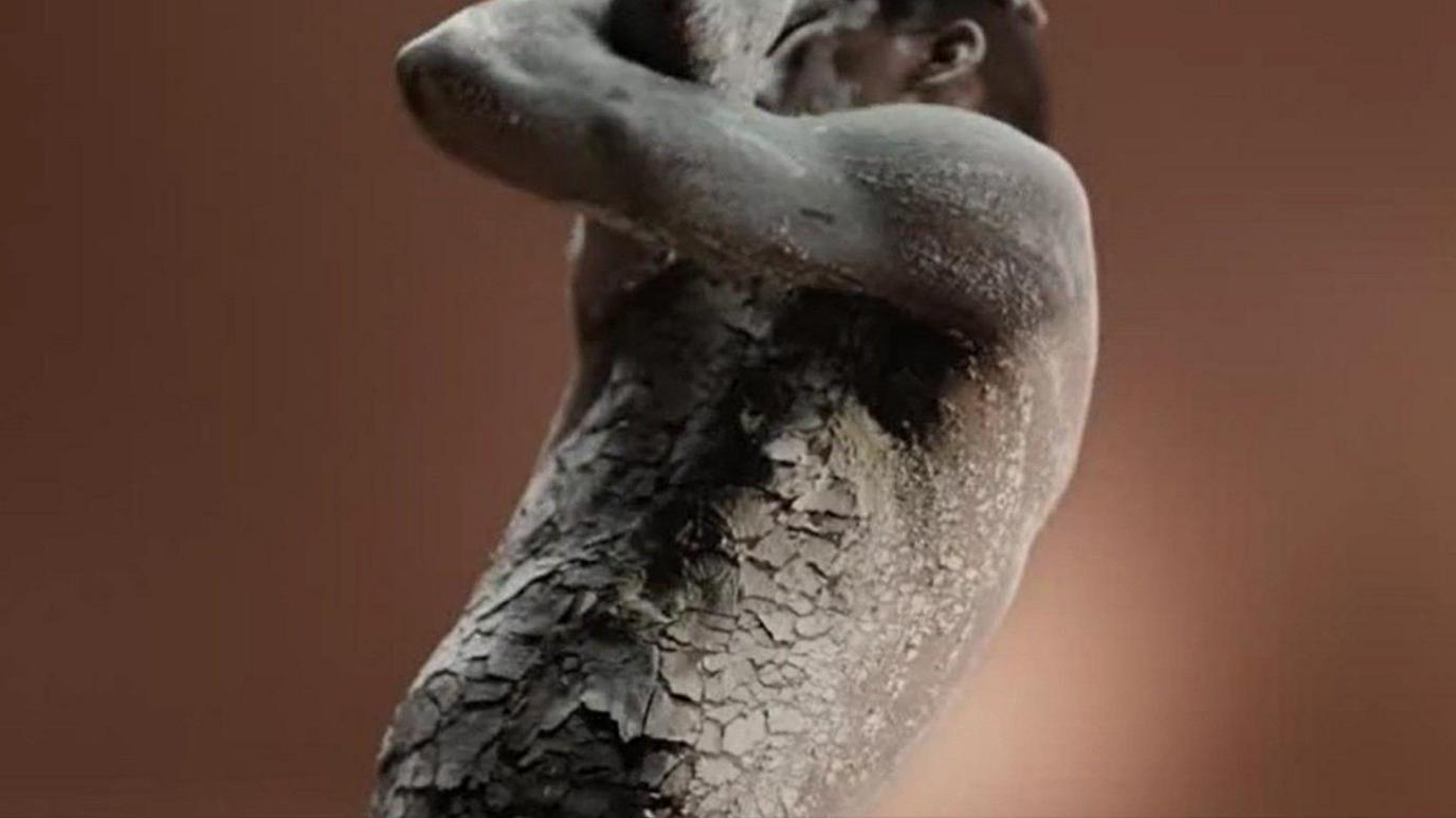 A topless man with black skin visible from his torso up covered in a grey clay-like material that is cracked. He is holding his arms across his face as though he is in pain.