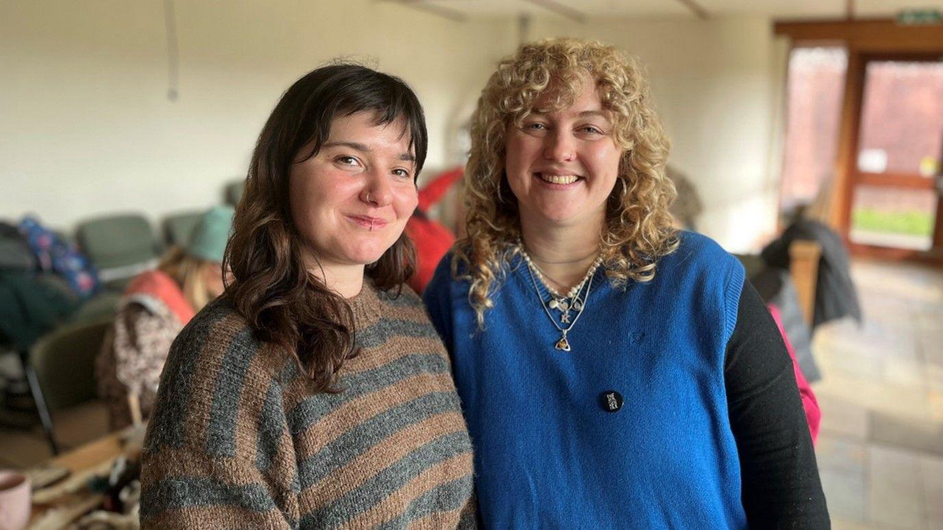 Meah Freeman-Burch (left) and Kat Cosgrove (right). Meah is wearing a brown and grey striped jumper and Kat is wearing a blue sweater vest with black undershirt. Meah has brown hair while Kat has blonde hair. 