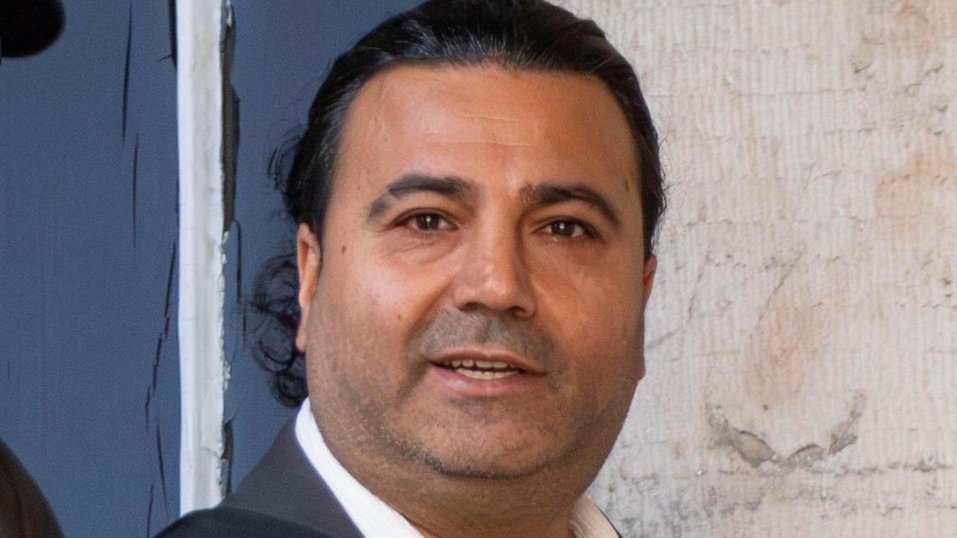 Kenan Baki looking straight at the camera. Only his face is visible. He has long dark hair and is clean shaven.