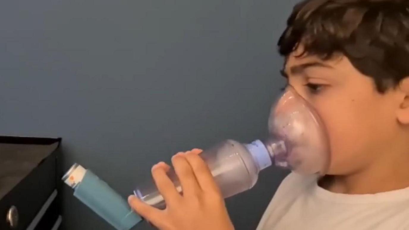 A young child holds an asthma inhaler system over his mouth and nose. There is an air-tight plastic mask connected to a tube which fits over his mouth. This in turn is connected to the inhaler.