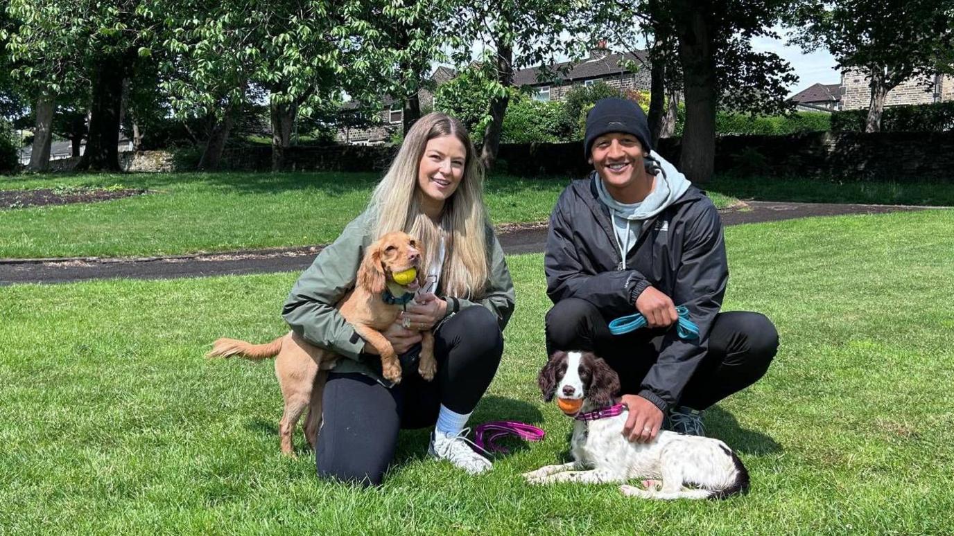 A woman and man kneeling on grass in a park with two spaniel dogs