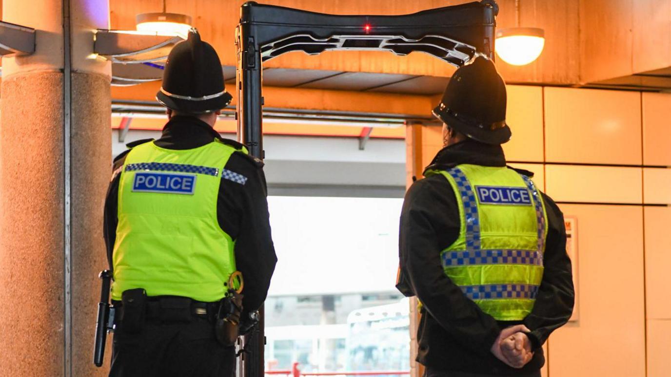 Two police officers standing in front of a knife detector arch at Sunderland's Park Lane Metro station. Their backs are turned to the camera. They are wearing black clothing, high visibility vests and black helmets.