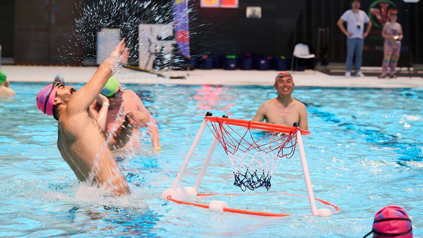 People standing in a swimming pool throwing balls in the air. There is an red, white and black net floating in the pool