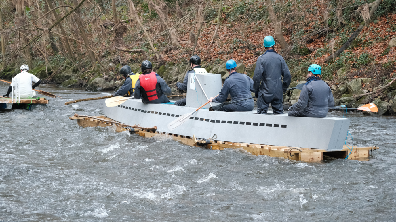 A raft shaped like a grey submarine with six people sat on top of it holding oars.