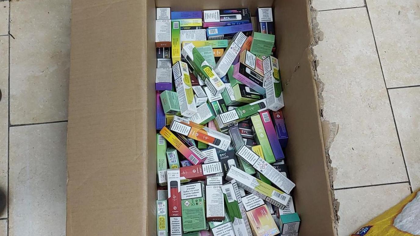 A box full of illegal vapes, set on the ground. Part of the box lid has been broken off.