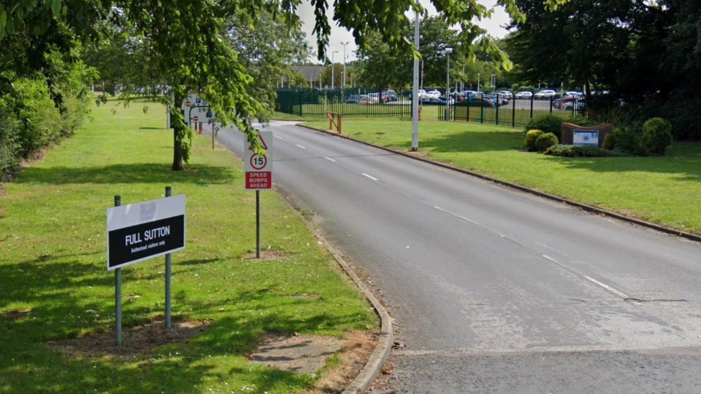 The entrance to HMP Full Sutton. A black and white sign stands on a neatly cut grass verge beside a roadway, with fencing and a car park in the background.