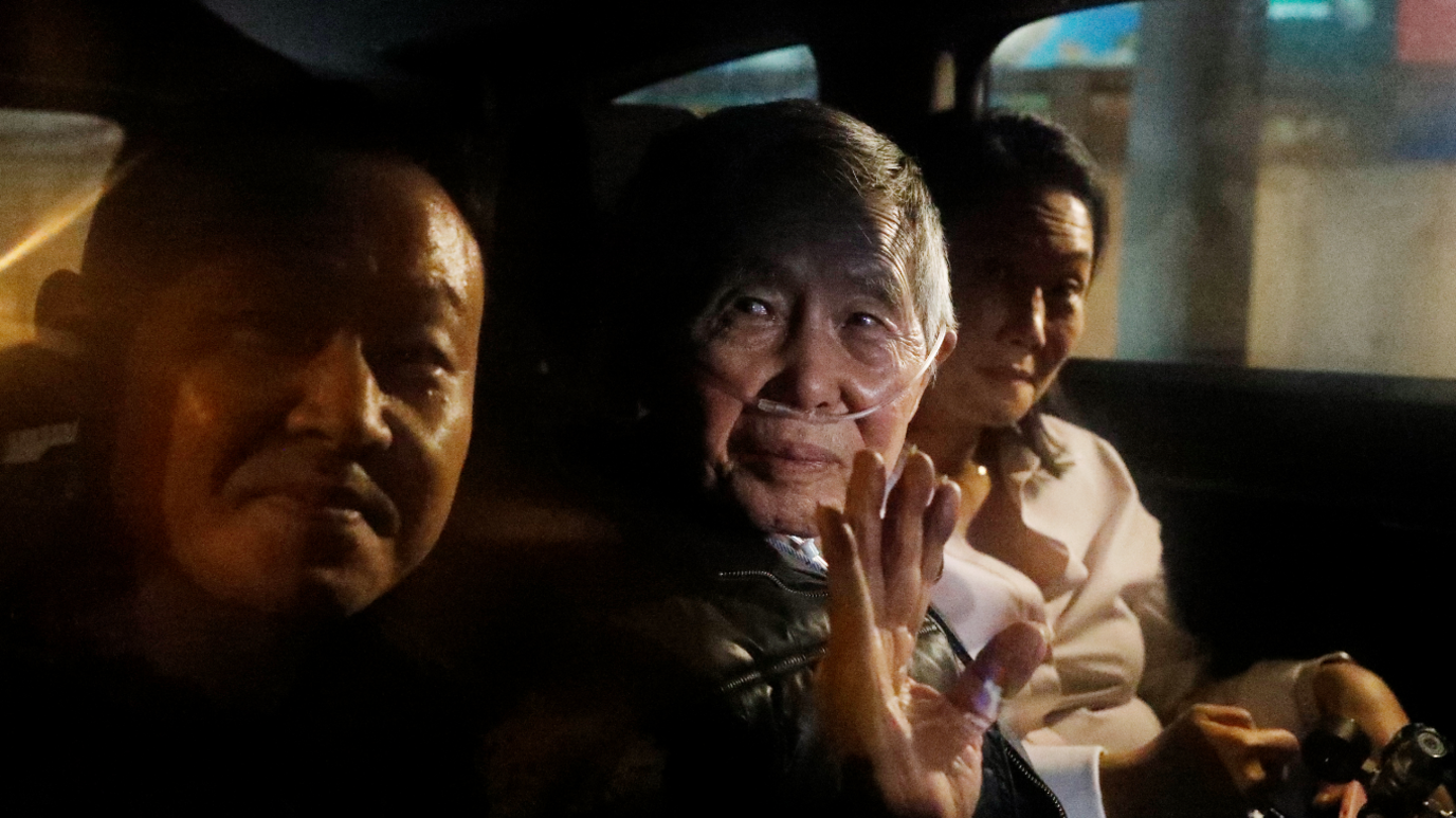 Former Peruvian president Alberto Fujimori sits in the middle seat at the back of a vehicle, waving, as he leaves Lima's Barbadillo prison. His daughter Keikio is sat next to him wearing white and another male is to Fujimori's right. Fujimori has a supply of oxygen running through his nostrils.