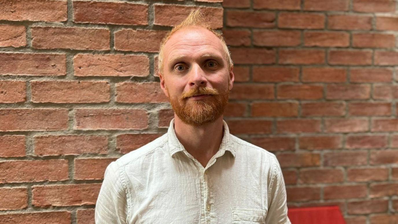 Martin smiling. He has ginger hair and a beard. He is wearing a cream shirt. The background is a red brick wall.