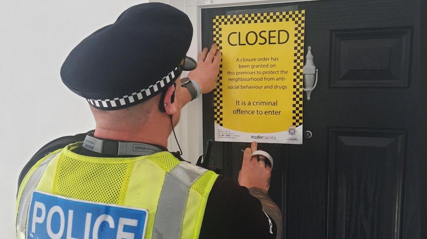 A police man wearing a green high visibility jacket pasting a yellow closure notice on a black door 