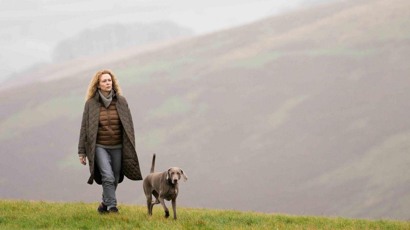 A woman in a long coat is walking her grey dog along a hill on Exmoor. It is foggy, lending the image a moody quality.