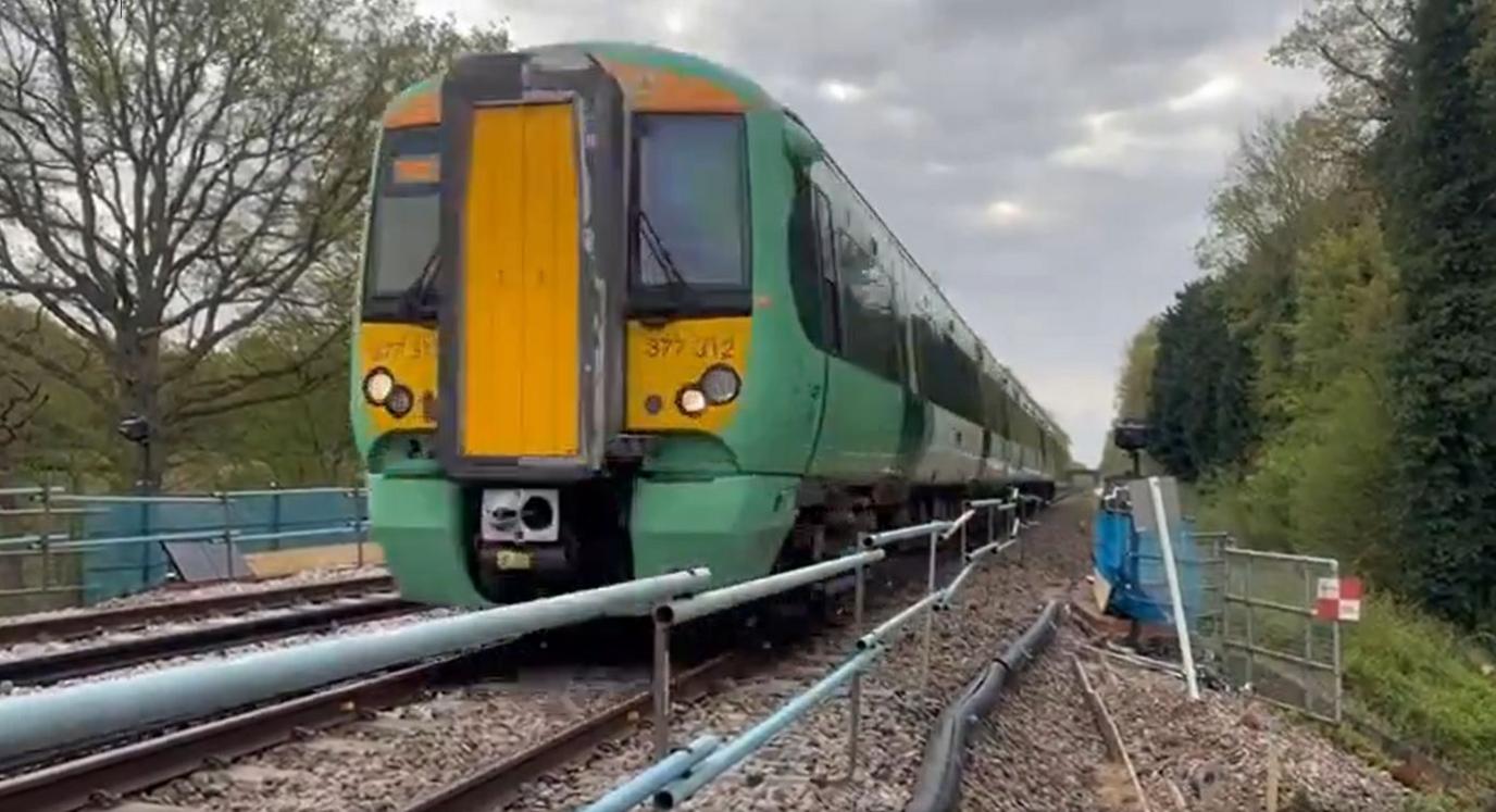 A green and yellow train is pictured moving along a railway track with temporary sidings and construction equipment visible nearby 