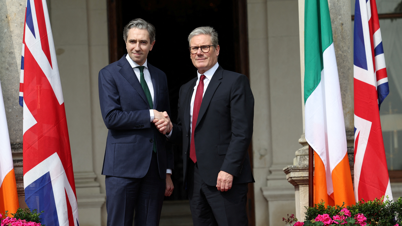 Taoiseach Simon Harris shaking hands with Prime Minister Sir Keir Starmer outside Farmleigh House in Dublin. There are Irish and Union Jack flags on either side of the two leaders. 