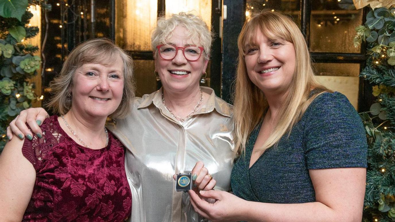 Three women posing for a photo following the award show. The woman on the left has shoulder length brown hair with grey streaks, and is wearing a red lace blouse. In the middle is Ann Remmers, with short white curly hair, wearing a silver button up blouse and red framed glasses. She is smiling and holding the award, which is a small metal coin in a plastic square case. On the right is another woman with long blonde hair, wearing a blue sparkly top . 