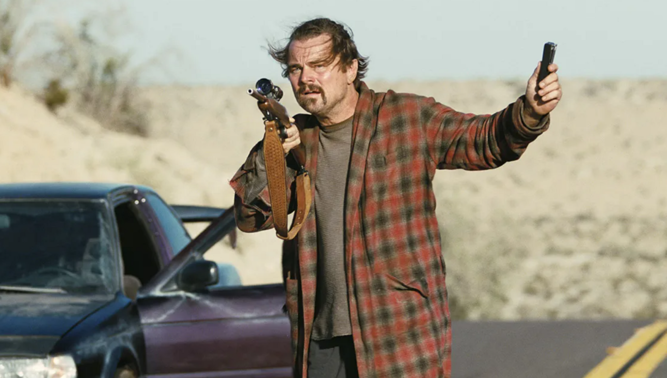 Leonardo DiCaprio as Bob in One Battle After Another standing outside a car holding a gun 