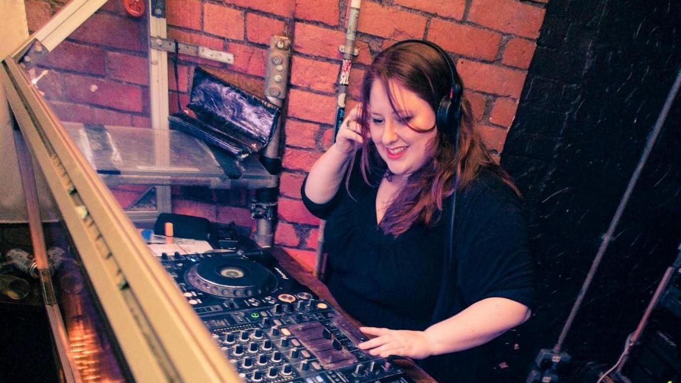 Esyllt Williams, menyw gwallt brown hir gyda mewn crys-t du a chlust-ffonau mewn bwth DJ