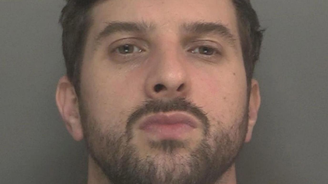 A mugshot of Thomas Cashman, who has dark brown hair and black stubble, and purses his lips at the camera with a defiant expression