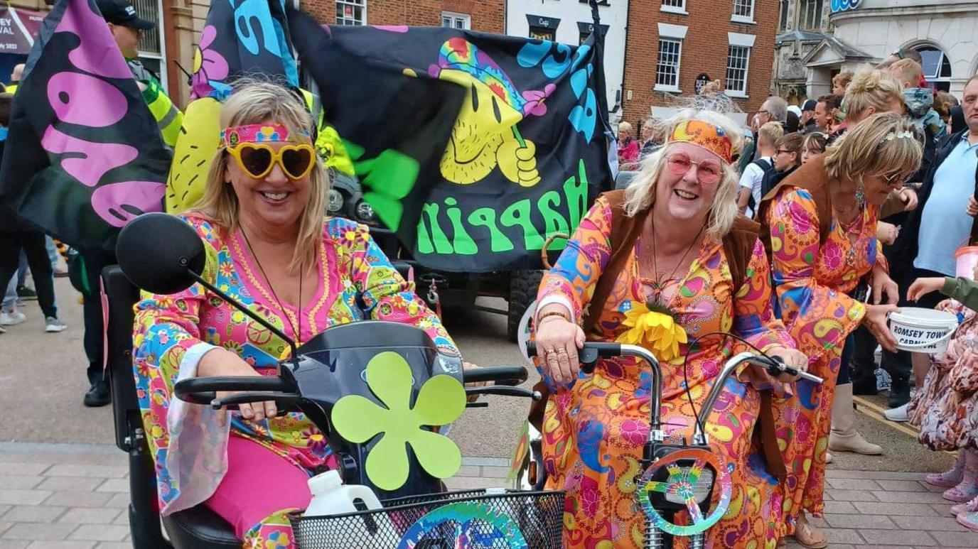 Two women on mobility scooters are dressed in 70s-themed hippie fancy dress. In the background there are large black flags with colourful words written on them.