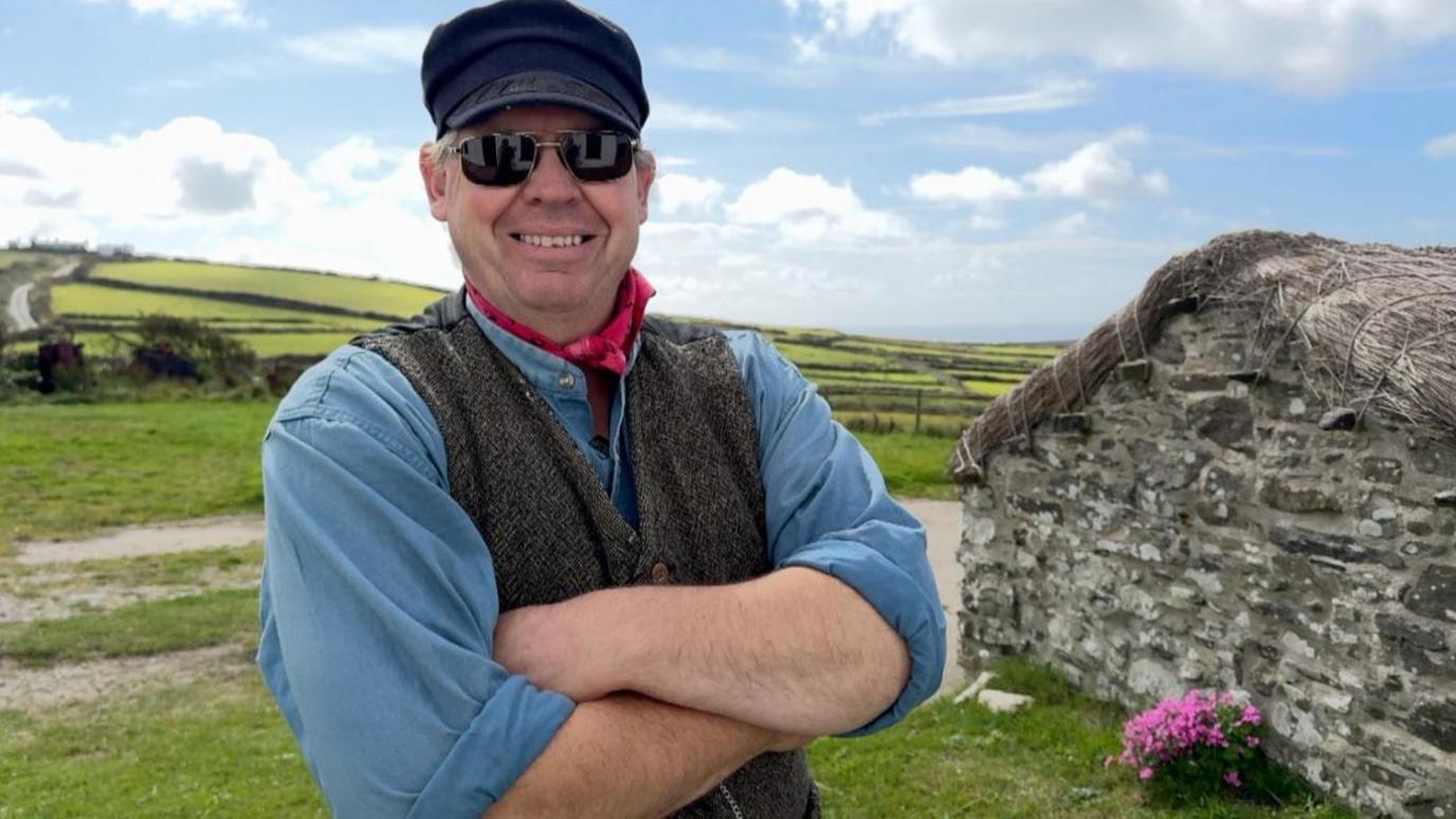Pete Kelly is wearing a blue shirt under a brown waist coat, and red cravat. He's smiling, wearing sunglasses and a flat cap. He's standing in front of rolling green hills and a small stone building with a thatched roof to the right.