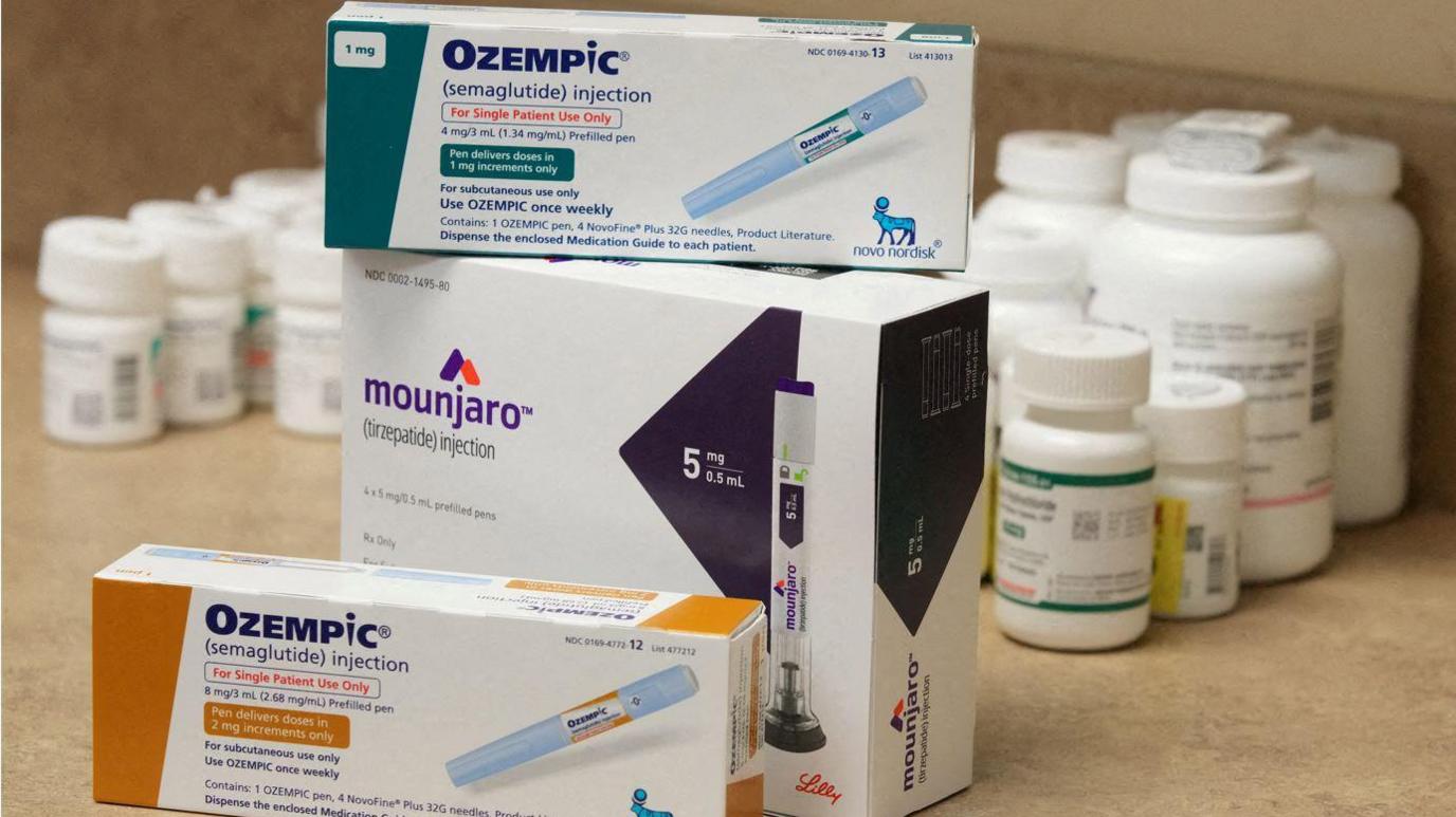 Boxes of Ozempic and Mounjaro injectables in cardboard boxes on a table with pill boxes seen in the background.