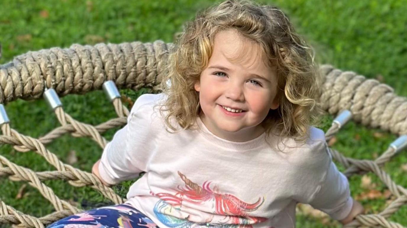Amber is sitting on a wide circular rope seat which has rubber matting below it and grass beyond it. She is smiling at the camera. Her arms are outstretched behind her and her legs are crossed in front of her. She has shoulder-length blonde curly hair and clue eyes. She is wearing a brightly coloured sweatshirt with a unicorn on it, unicorn-printed leggings and unicorn-printed wellies.