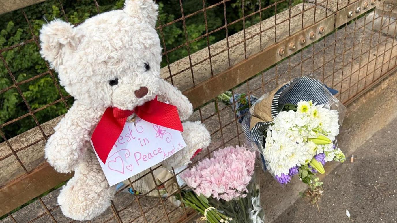A teddy bear and two bouquets of flowers laid at a fence.