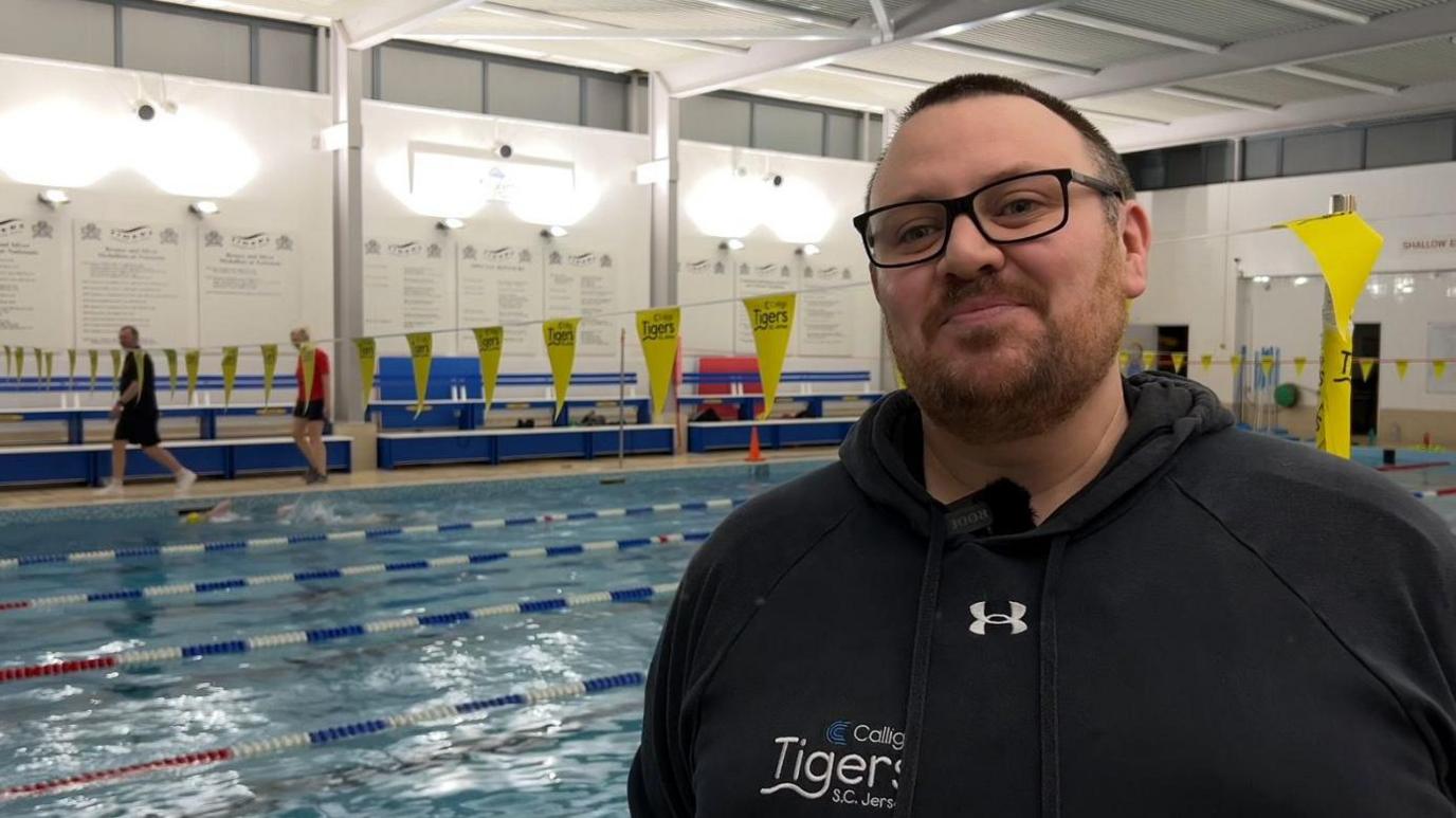 Nathan Jegou, a man with short brown hair, a mid-length brown beard and dark rimmed glasses. He is wearing a dark hooded sweatshirt with white writing on the front. He is standing in front of a swimming pool, with lane markings. Yellow flags stretch across the pool behind him.