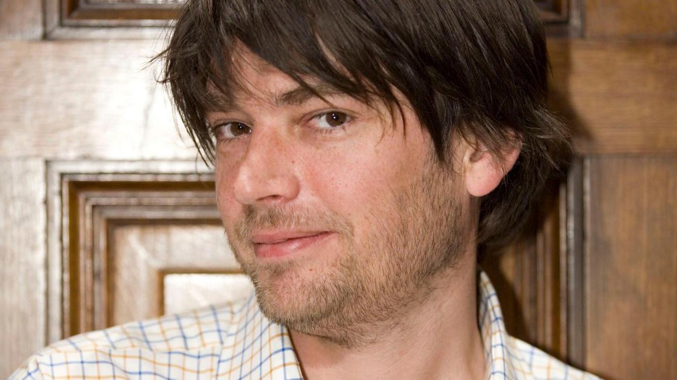 Alex James looking sideways at the camera and smiling with his lips closed. He has brown eyes and shaggy brown hair with a long fringe and it is just below his ears at the back. He has a short beard and is wearing a shirt with a blue and yellow check.