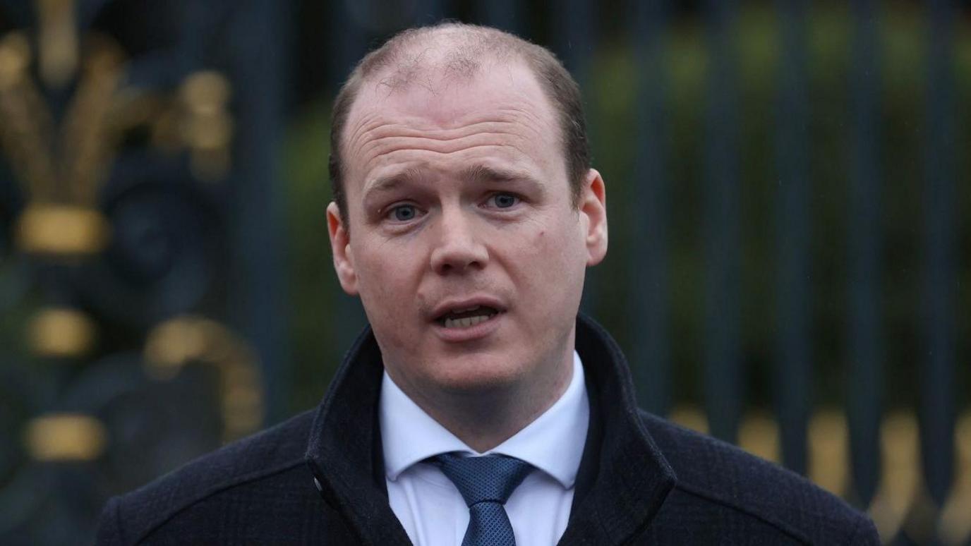 Gordon Lyons who is balding, speaking outdoors wearing a dark coat, white shirt and tie