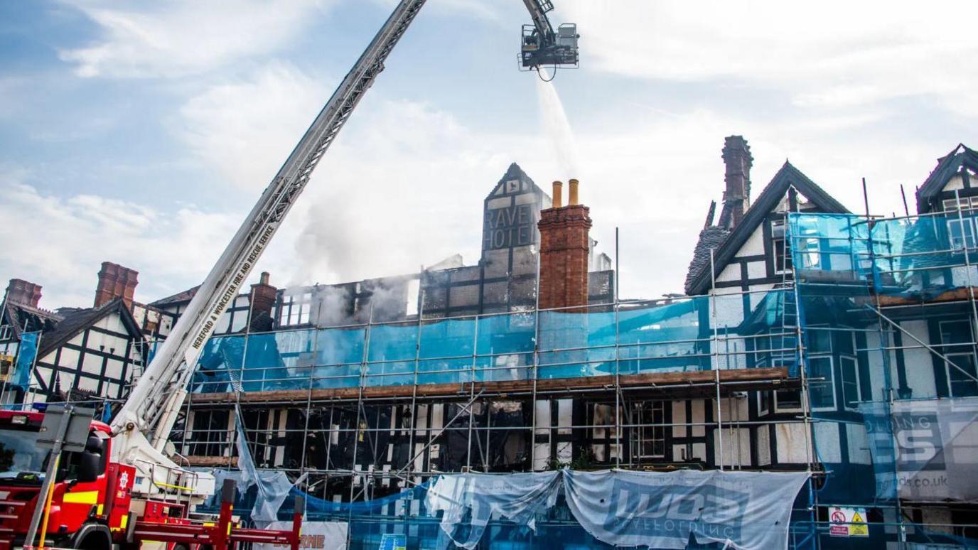 A black and white timbered building covered in scaffolding, with a fire engine and crane squirting water from above. The name Raven Hotel can be seen on a tall chimney.