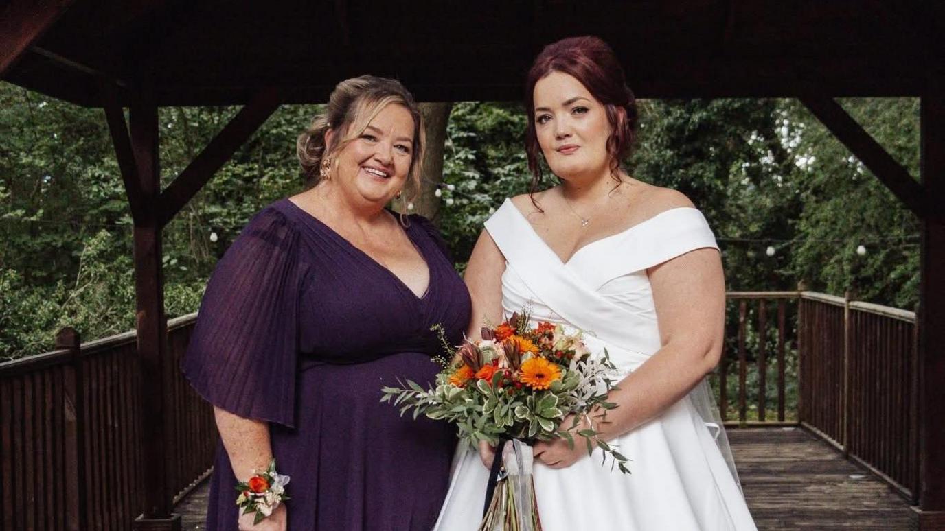 A woman in a purple dress stands next to another woman in a white wedding dress holding a bouquet of flowers. 