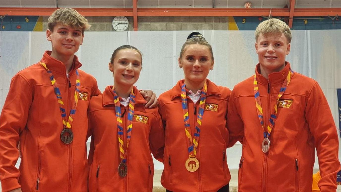 Four young Manx athletes, two women and two men, wear red jackets and stand with arms around each other. They each have different coloured medals around their necks.