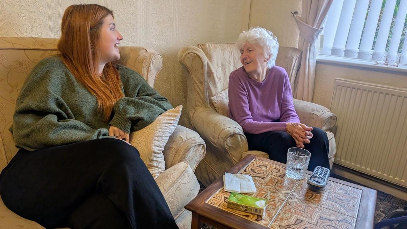 Two women are seated and engaged in conversation. On the left, a younger woman with long, red hair wears a green jumper and black trousers. Opposite her, an older woman with short white hair wears a purple sweater and black trousers. In front of them is a coffee table with a glass of water and remote control on. 
