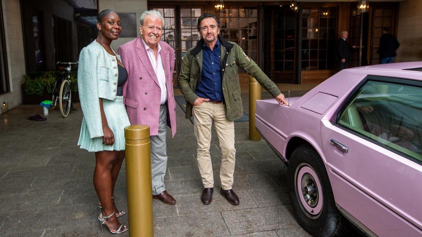 The owners of the Aston Martin, Emma and Roger Dudding, stand with Richard Hammond next to the pink Aston Martin, which is parked in front of a hotel. She is wearing a light blue skirt and jacket and he has a pink suit jacket and grey trousers. Richard has light trousers and black boots on and is also wearing a green waxed jacket.