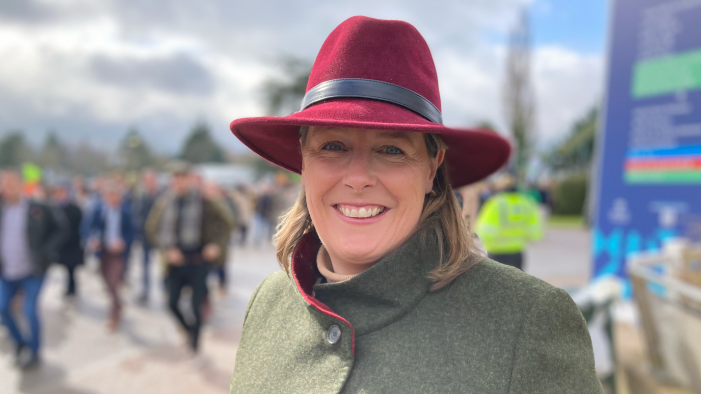 Gemma Steve wearing a wine-red hat and green coat with racegoers walking behind her.