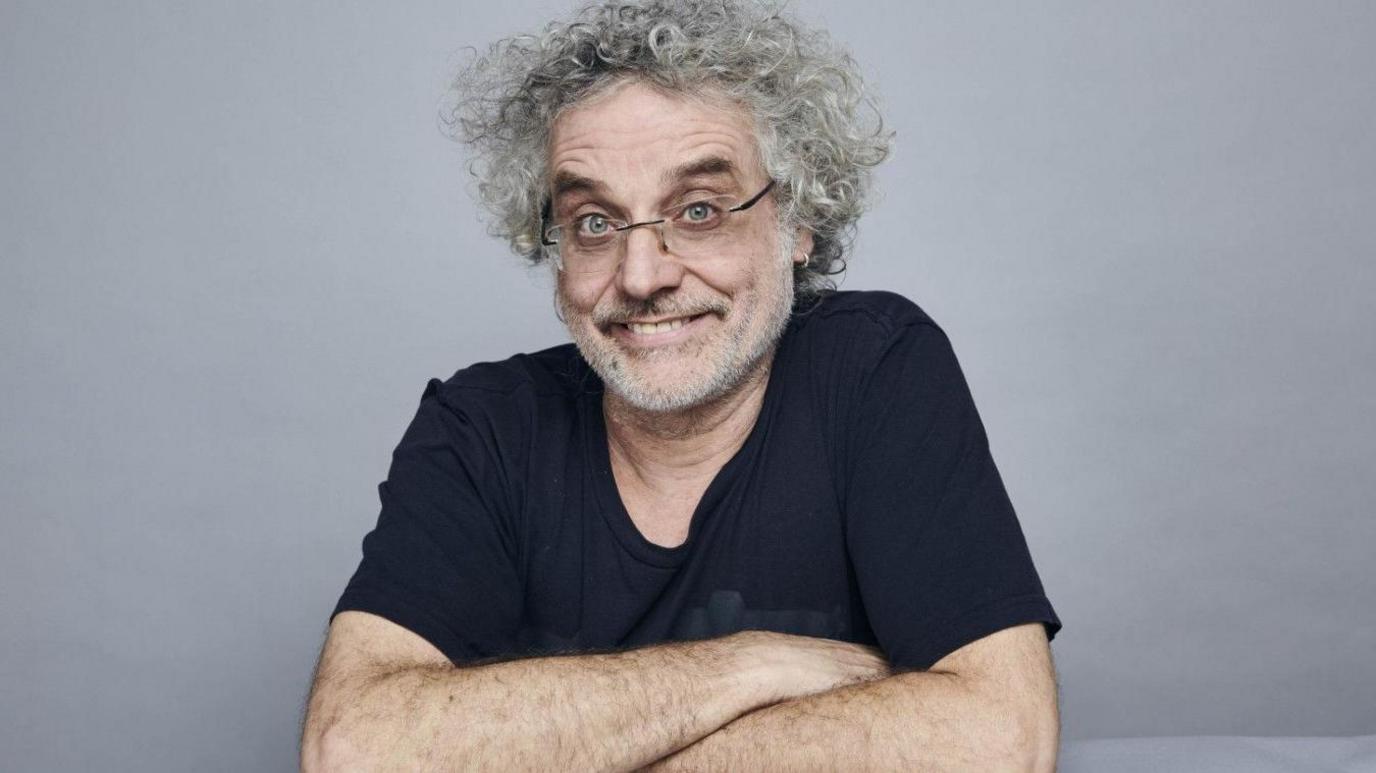 Studio picture of Matt Perry - he has curly grey hair and glasses, smiling with arms crossed in front of him. He is wearing a blue t-shirt and has a small hoop earring in his left ear.