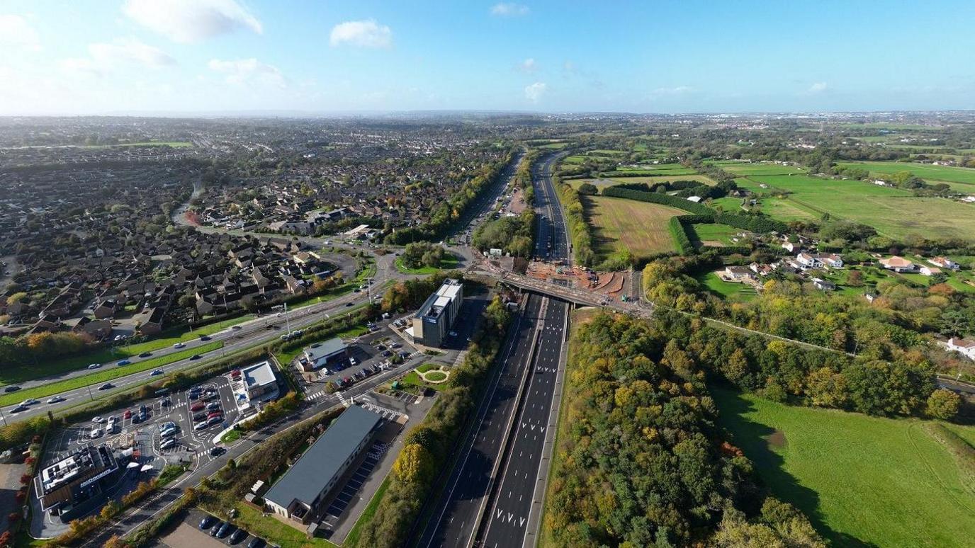 A drone image shpws the M4 motorway close to Bristol with no traffic in one carriageway due to roadworks. Also visible is the Badminton Road Bridge where the work is taking place, with vehicles and large amounts of earth piled on the road surface. It is a sunny day with blue sky and the city of Bristol and surrounding countryside is also visible