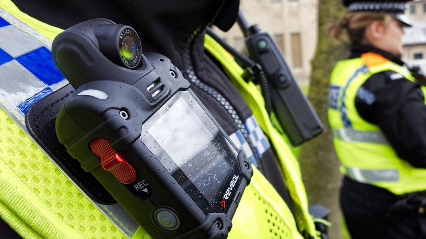 The image shows a close-up of a small black box attached to the hi-vis vest of a police officer. A red button on the side has the letters "REC" in white underneath.