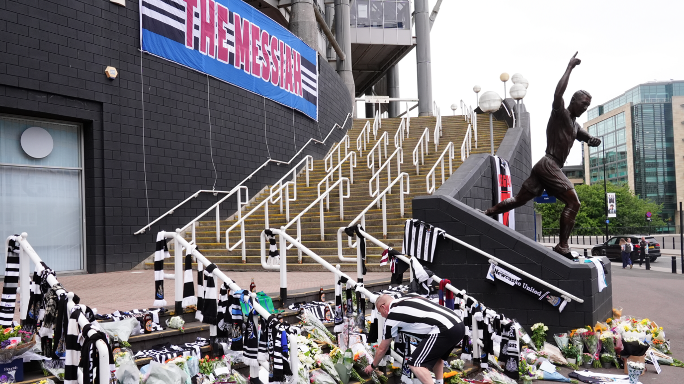 A man in a Newcastle United shirt places a bouquet on the steps St James' Park, which are covered by flowers and NUFC shirts and scarves.  A flag reading The Messiah - one of the nicknames fans had for Keegan - is attached to the wall of the stadium. 