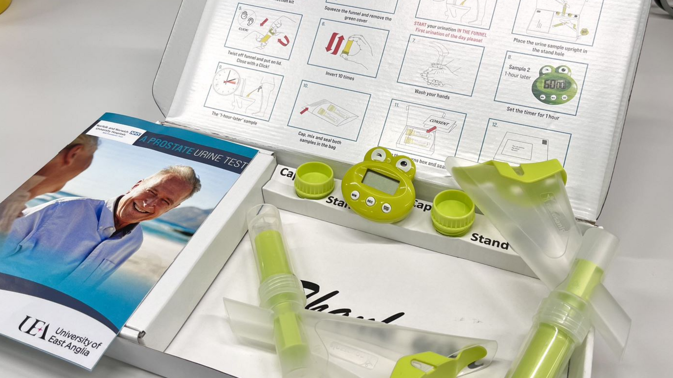 An open shallow white box containing green and clear plastic items of testing kit and a leaflet called A Prostate Urine Test, on the cover of which is a man in his 60s smiling at a woman and the words UEA University of East Anglia