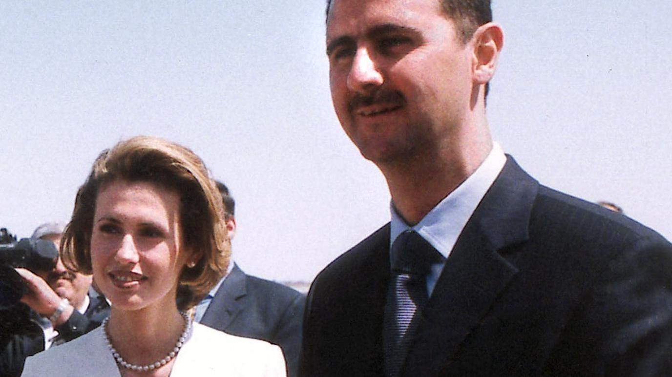 An old blurry picture of Asma al-Assad in a pearl necklace and white jacket and Bashar al-Assad in a dark suit, both slightly squinting outside