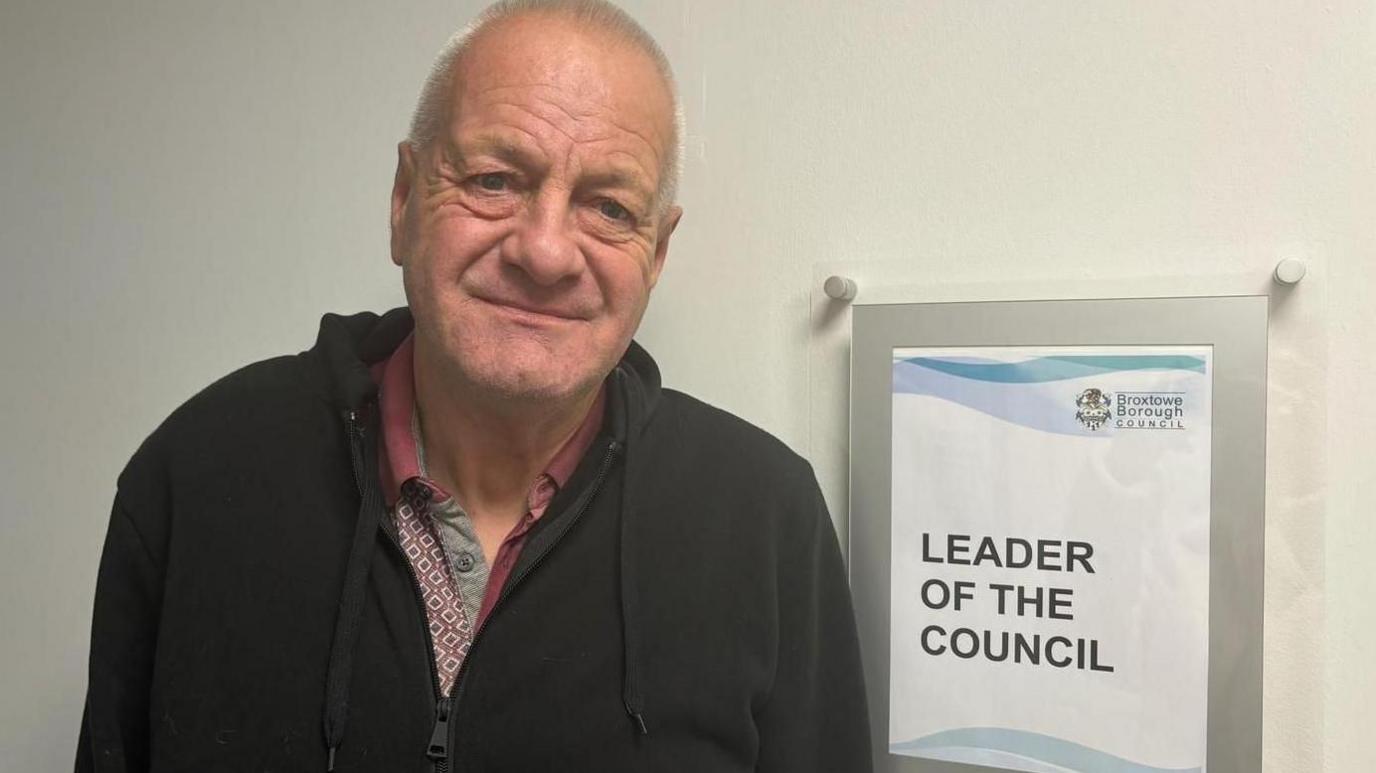 Milan Radulovic wearing a black hoodie and red collared shirt in front of a sign which says leader of the council