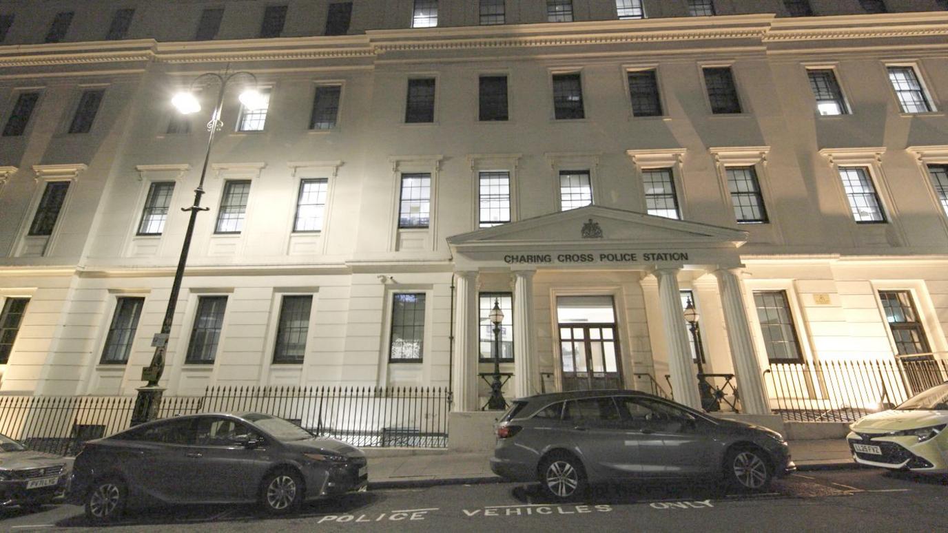 An exterior view of Charing Cross Police Station at night: a white stucco neo-classical building with a four-pillared portico over the entrance and the Met Police's crest above the door.