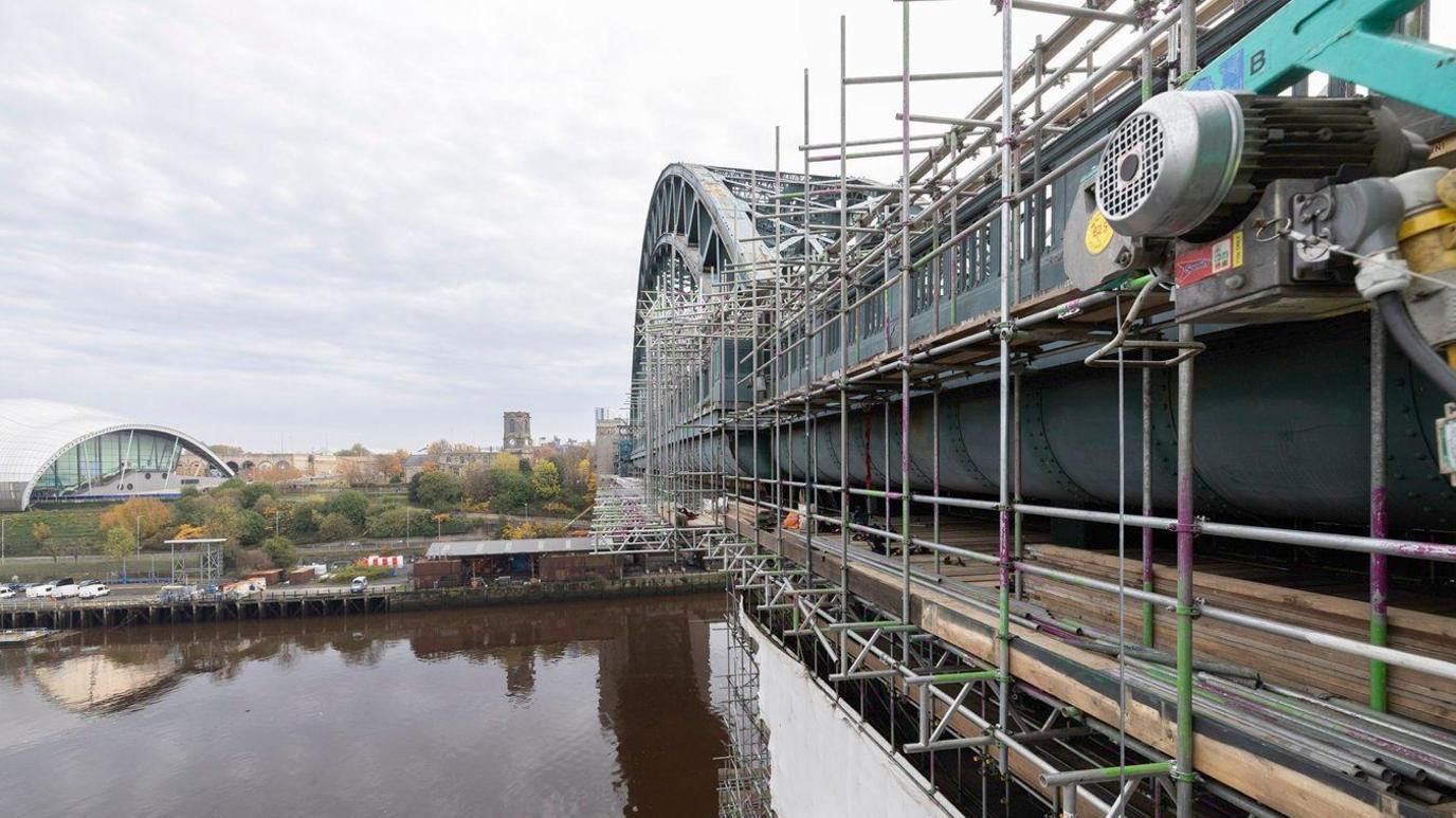 To the right, there is a large green metal arched bridge over the river, with scaffolding on it. In the distance there are trees and buildings and a car park. It is a cloudy day.