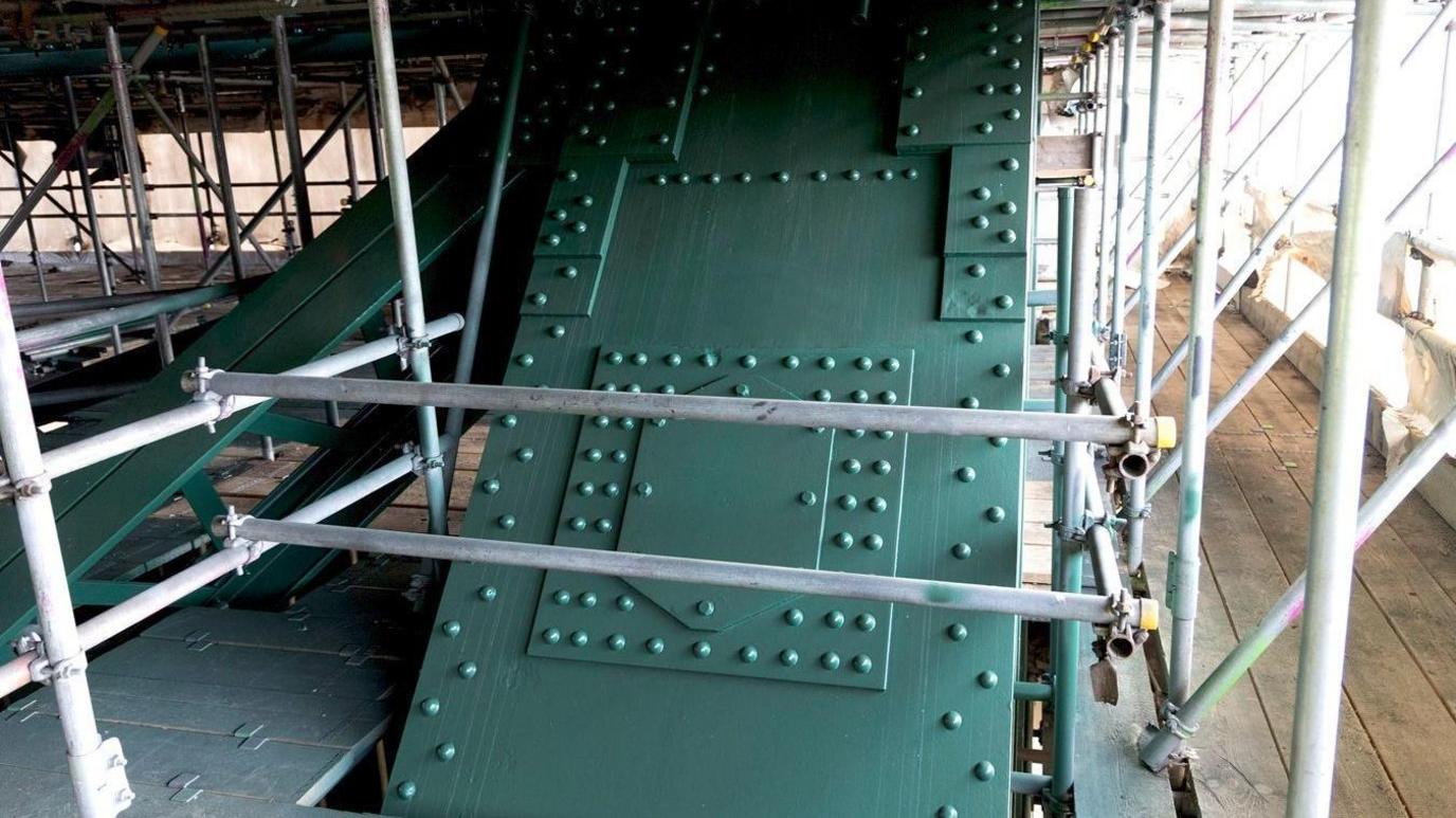 Part of the green, freshly-painted bridge behind grey scaffolding. 