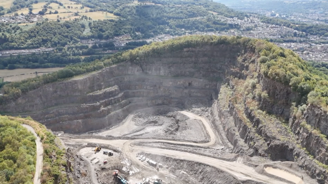 In the foreground is the quarry with lorry tracks and the exposed rock face of the mountain. Beyond that is a view down the valley with trees, fields and villages in the distance.